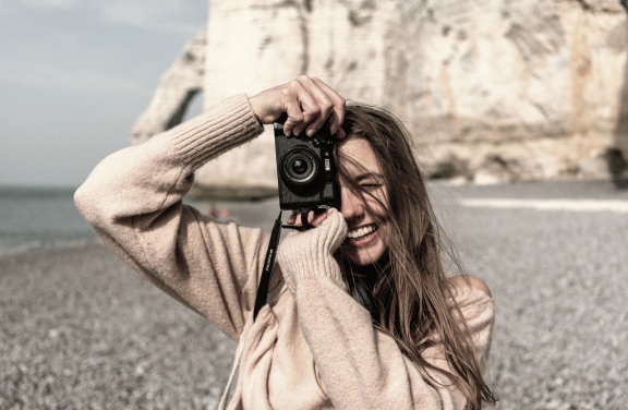 Smiling woman on a rocky beach in the Lake Constance region, holding a camera to her face, with cliffs in the background.