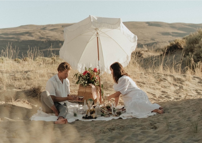 Ein Paar genießt ein Picknick unter einem weißen Sonnenschirm auf sandigem Boden mit Hügeln im Hintergrund und fängt die Romantik eines Hochzeitswochenendes in Südtirol ein.