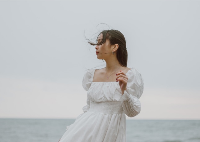 Eine Frau in einem weißen Kleid steht an einem wolkenverhangenen Tag am Meer, der Wind weht ihr Haar - ein heiterer Moment, der an ein Hochzeitswochenende in der malerischen Bodenseeregion erinnert.