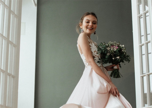 Eine lächelnde Frau in einem weißen Kleid mit einem Blumenstrauß in der Hand, die in einem Innenraum an großen Fenstern steht und den Charme der Bodensee-Region ausstrahlt.
