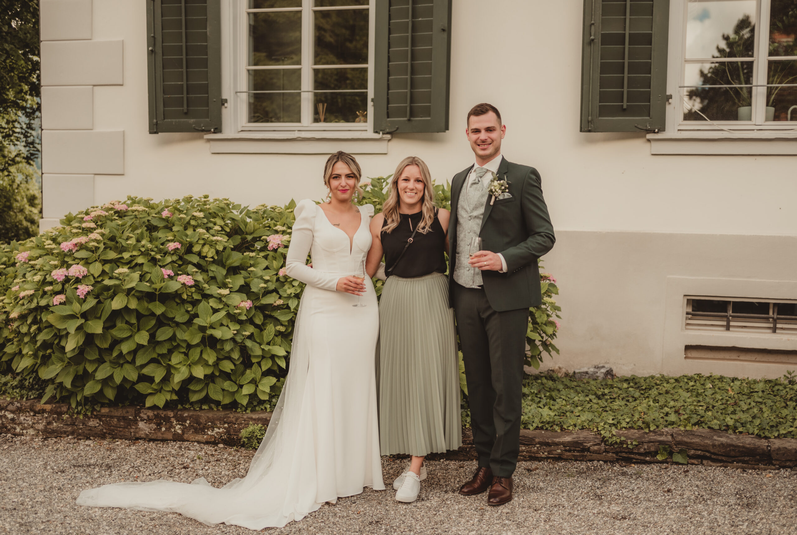 Braut, Bräutigam und ein Gast stehen lächelnd im Freien vor einem Haus mit grünen Fensterläden und Hortensiensträuchern und fangen den Charme der Bodensee-Region ein.