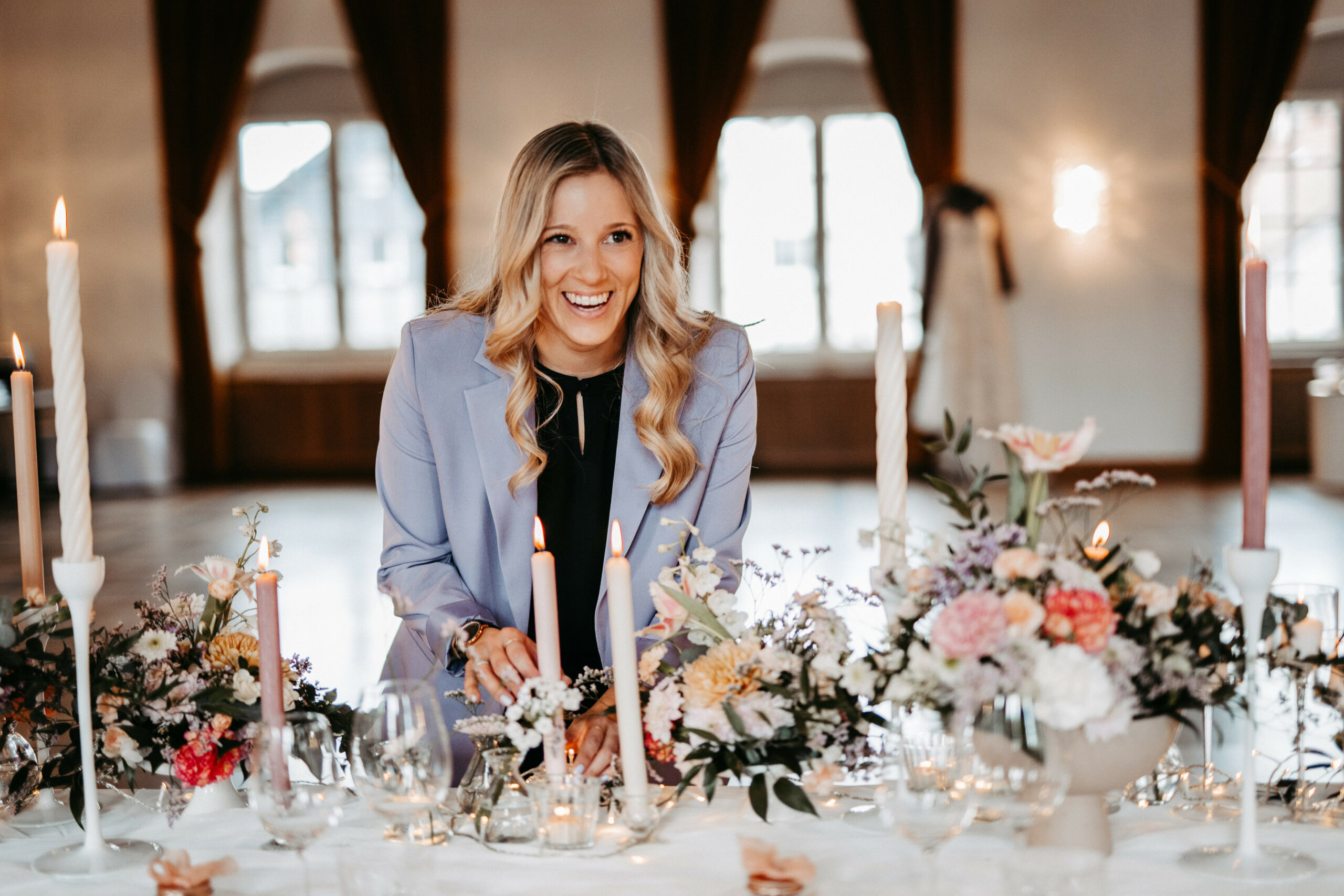 Eine lächelnde Frau im lavendelfarbenen Blazer arrangiert Blumen und Kerzen auf einem eleganten, geschmückten Tisch, inspiriert durch den raffinierten Stil der Bodensee-Region.