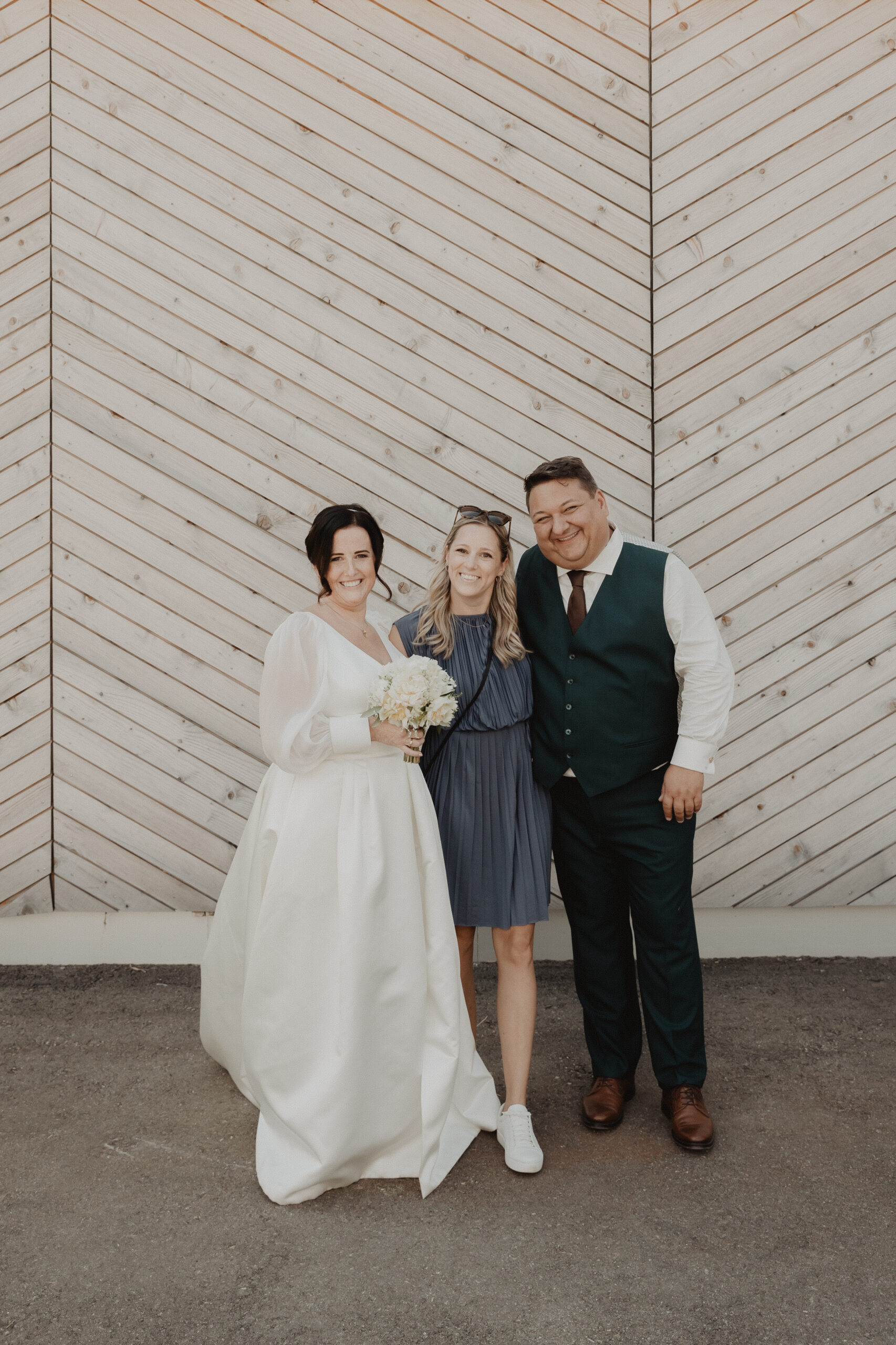 Braut und Bräutigam posieren und lächeln mit einem Gast vor einer hellen Holzwand im Chevron-Stil und fangen fröhliche Hochzeitserinnerungen ein, die von der Eleganz Österreichs inspiriert sind.