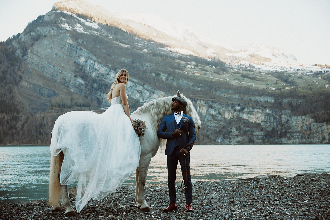Die Braut im weißen Kleid sitzt auf einem weißen Pferd neben dem Bräutigam im Anzug an einem See mit Bergen im Hintergrund und fängt die Romantik eines Hochzeitswochenendes in der schönen Bodenseeregion ein.