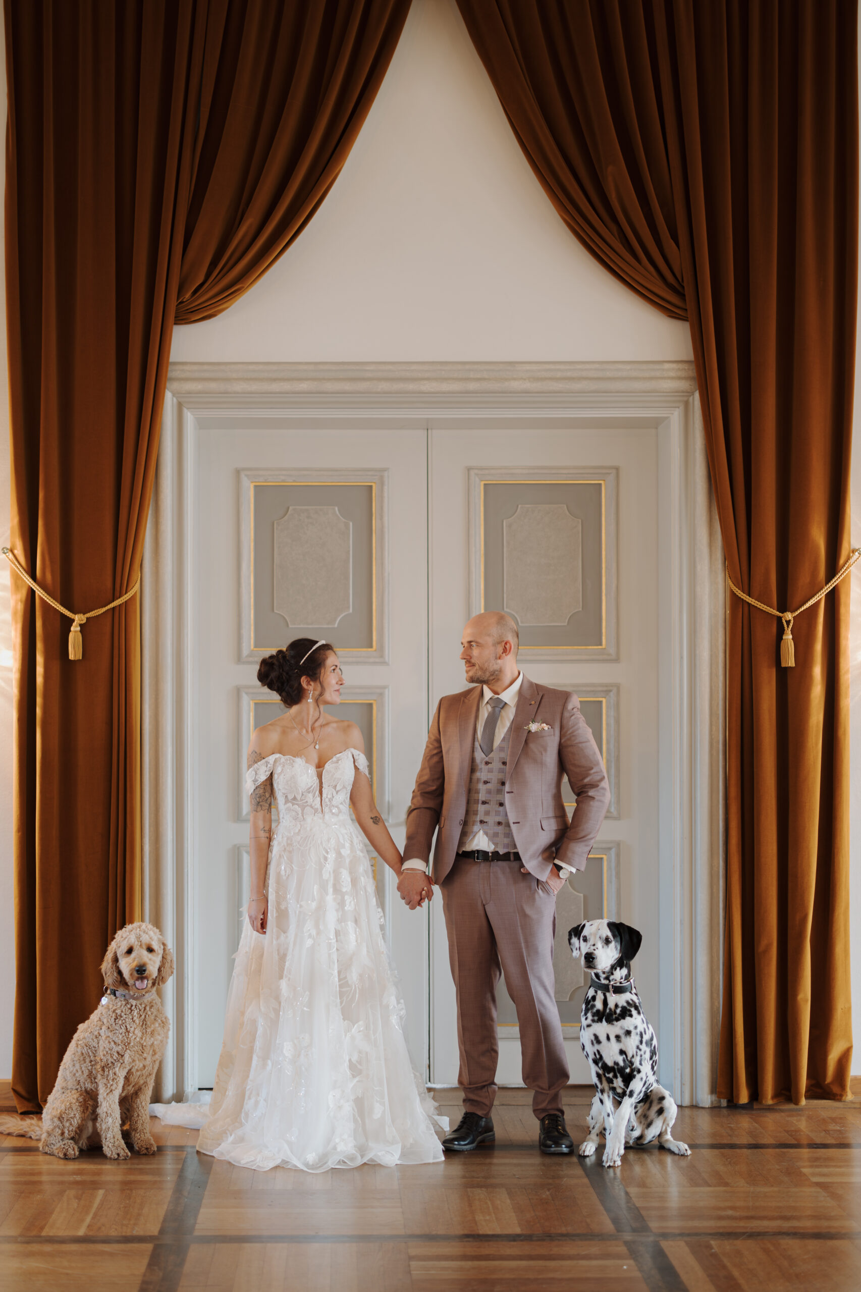Bride and groom holding hands, standing with a Labradoodle and a Dalmatine in front of elegant doors and curtains, capturing the charm of a wedding weekend in the Lake Constance region.