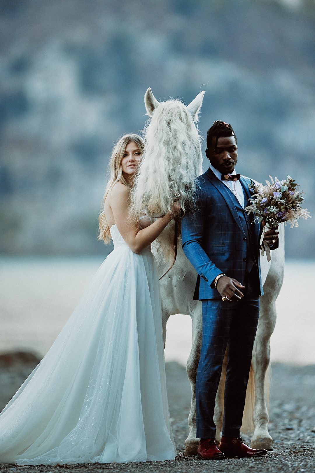 A bride and groom stand outdoors next to a white horse, with blurred mountains in the background, capturing the romantic charm of the South Tyrol region.