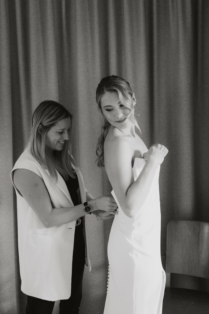 Eine Frau hilft einer Braut beim Anziehen ihres Kleides, während die Braut lächelt und nach unten schaut. Sie steht vor einem Vorhang in der schönen Bodenseeregion nahe Österreich und der Schweiz.