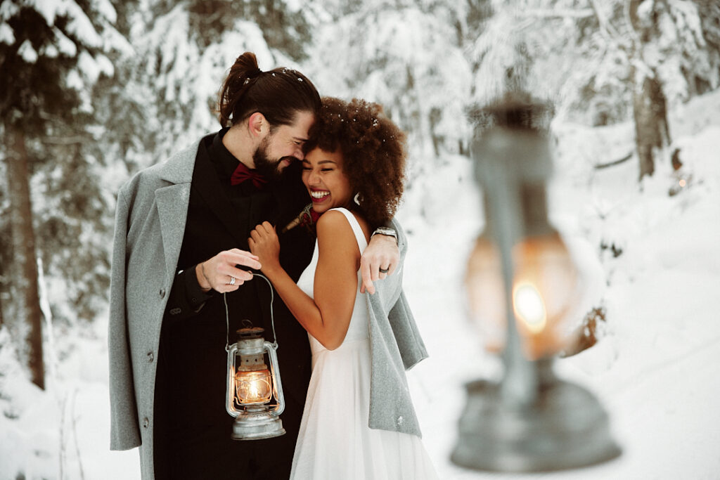 A couple in winter coats embrace in a snowy forest near the Lake Constance region, smiling and holding an old lantern.