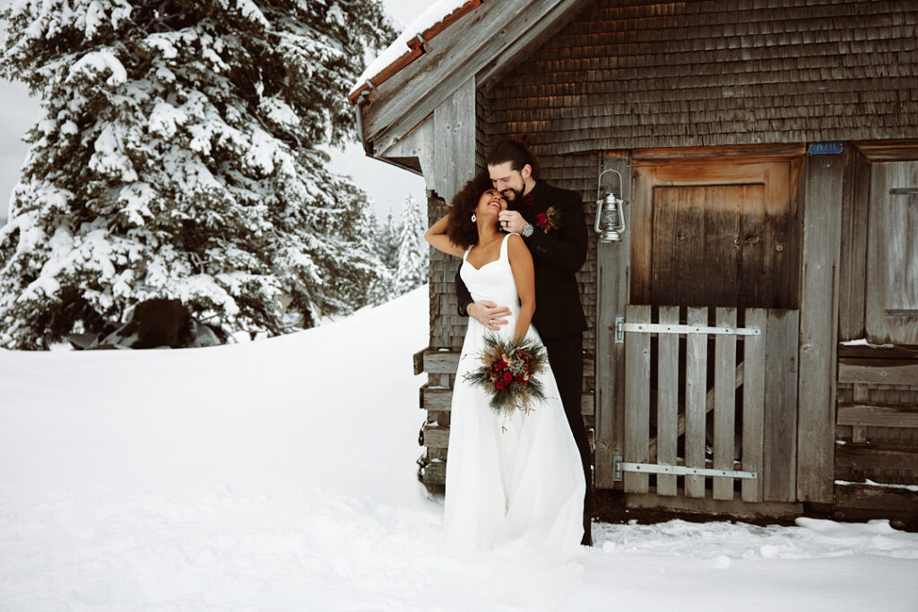 Bride and groom embrace and smile in front of a rustic wooden hut in a snowy winter landscape, capturing the magic of their wedding weekend in Austria.