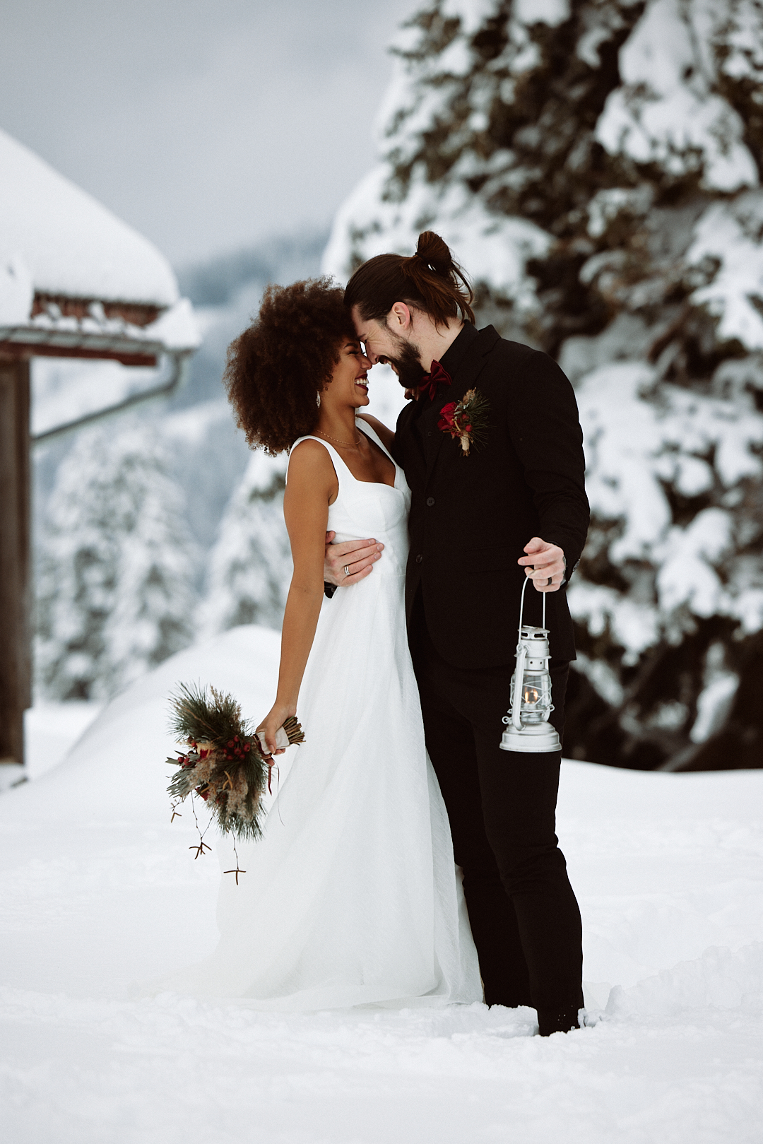 A bride and groom embrace outdoors in the snow in the Lake Constance region; she holds a bouquet of flowers, he holds a lantern, both are smiling warmly.