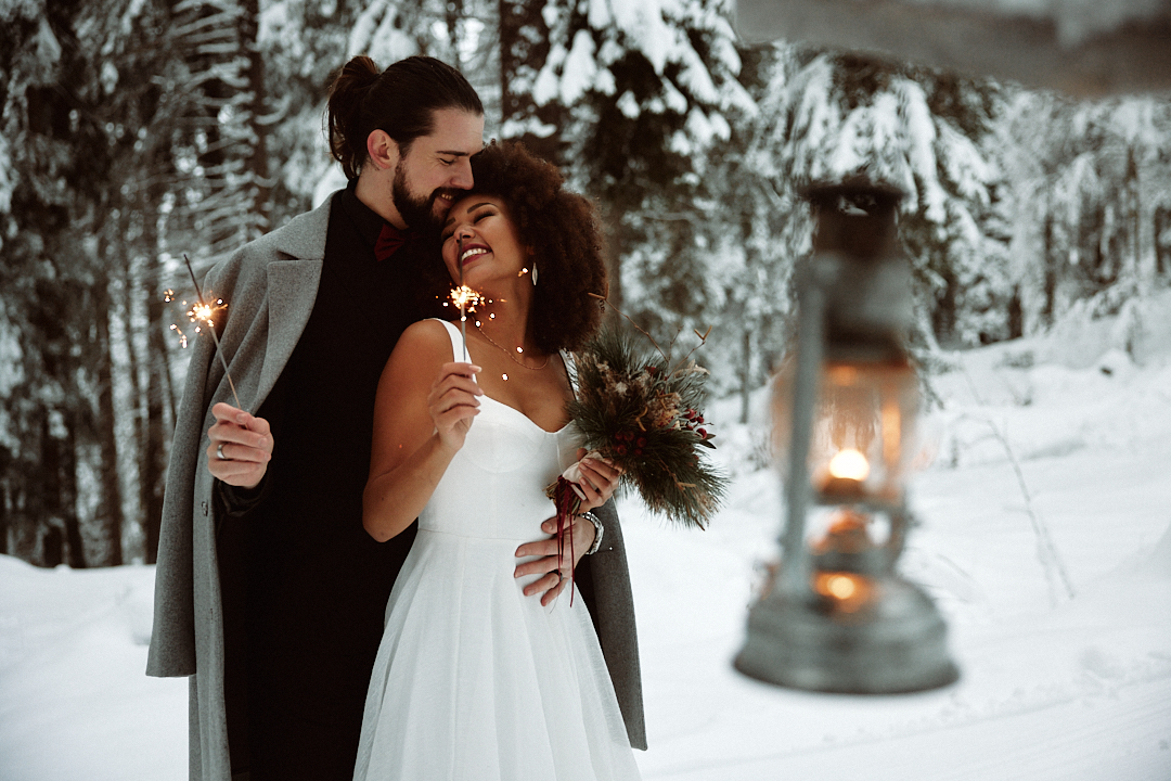 Ein Paar in Hochzeitskleidung hält lächelnd Wunderkerzen in einem verschneiten Wald in Südtirol, mit einer warm leuchtenden Laterne im Vordergrund.