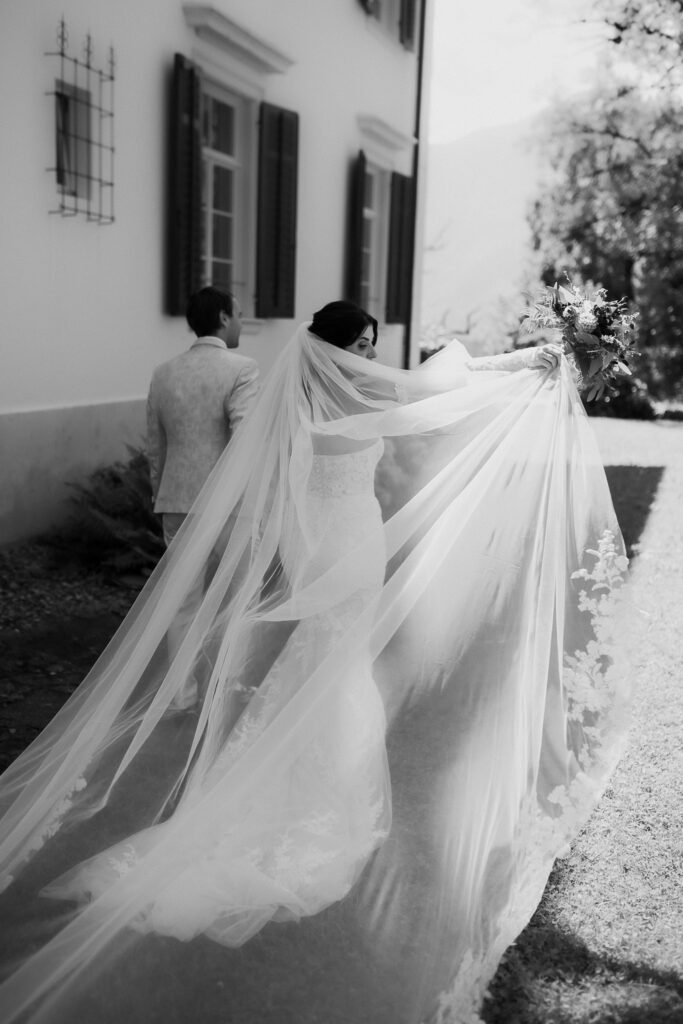 Eine Braut mit langem Schleier geht mit einem Mann im Freien spazieren, ihr Brautstrauß und ihr Schleier hängen im Sonnenlicht hinter ihr her und fangen die Romantik eines Hochzeitstages in der malerischen Bodensee-Region ein.