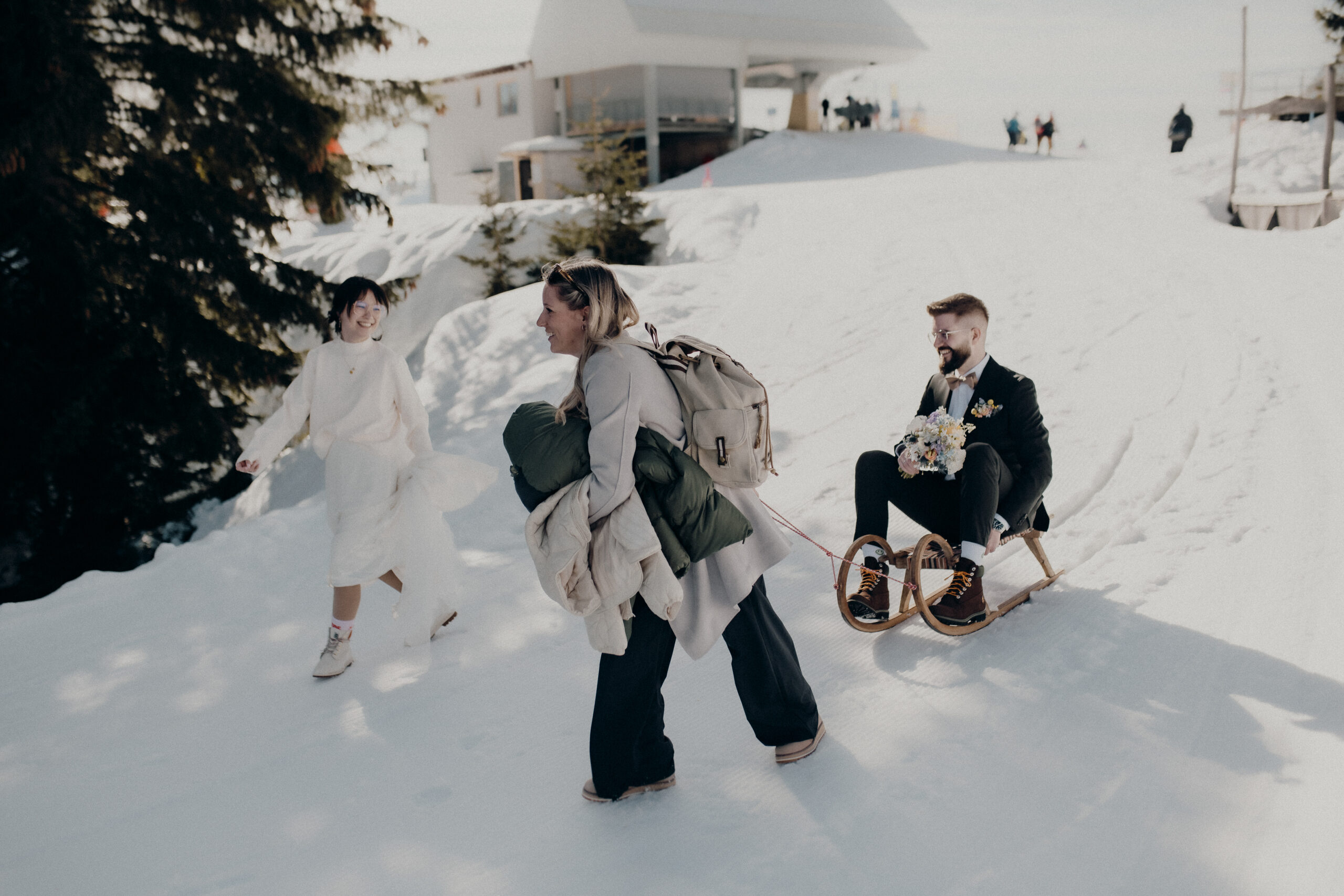 A woman pulls a man in a suit on a sledge while another woman walks next to them in the snow. They create happy memories of their wedding weekend in the picturesque Lake Constance region.