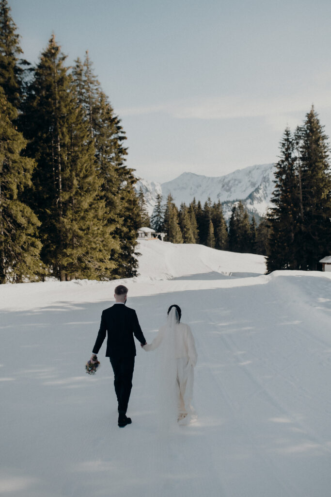 Bride and groom walk hand in hand across the snow-covered ground, surrounded by trees and mountains in the heart of South Tyrol.