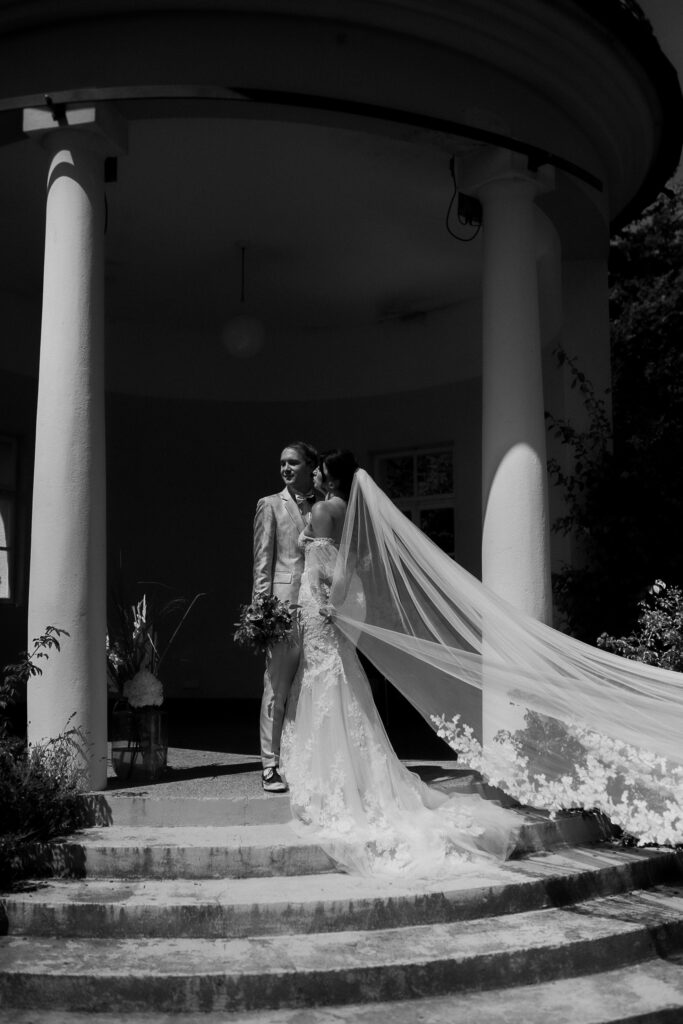 Eine Braut und ein Bräutigam stehen auf einer Treppe und umarmen sich, der Schleier der Braut weht hinter ihnen in der malerischen Bodensee-Region.