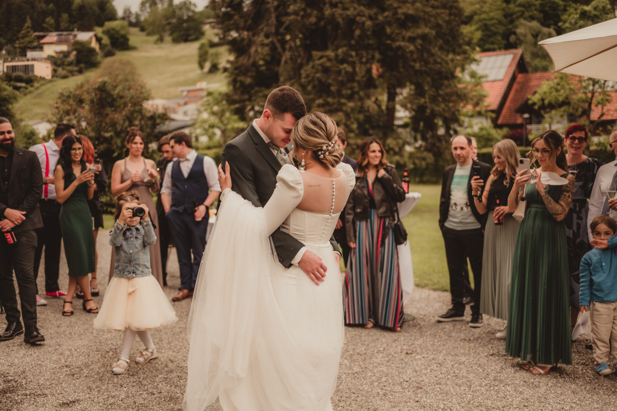 A bride and groom dance outdoors in the Lake Constance region, surrounded by smiling guests and lush vegetation.