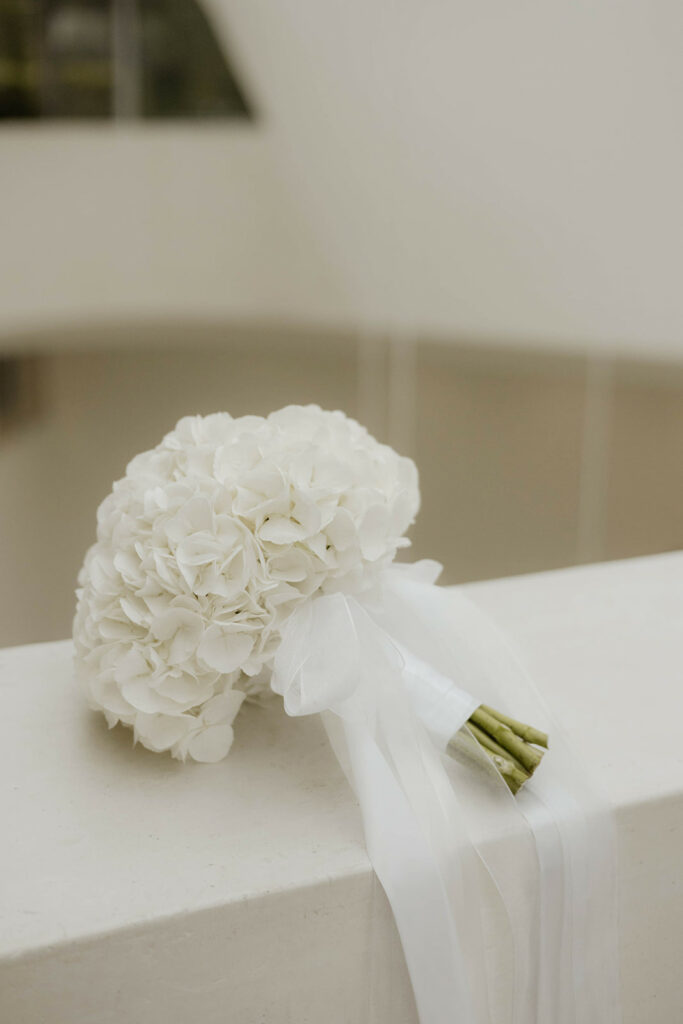 Ein Strauß weißer Hortensien mit weißer Schleife ruht auf einem weißen Sims, perfekt für ein Hochzeitswochenende in der Schweiz oder Österreich.