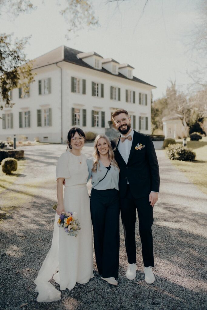Three people stand smiling in front of a large white house in the Lake Constance region; one is wearing a wedding dress, another a suit and the third casual clothes.