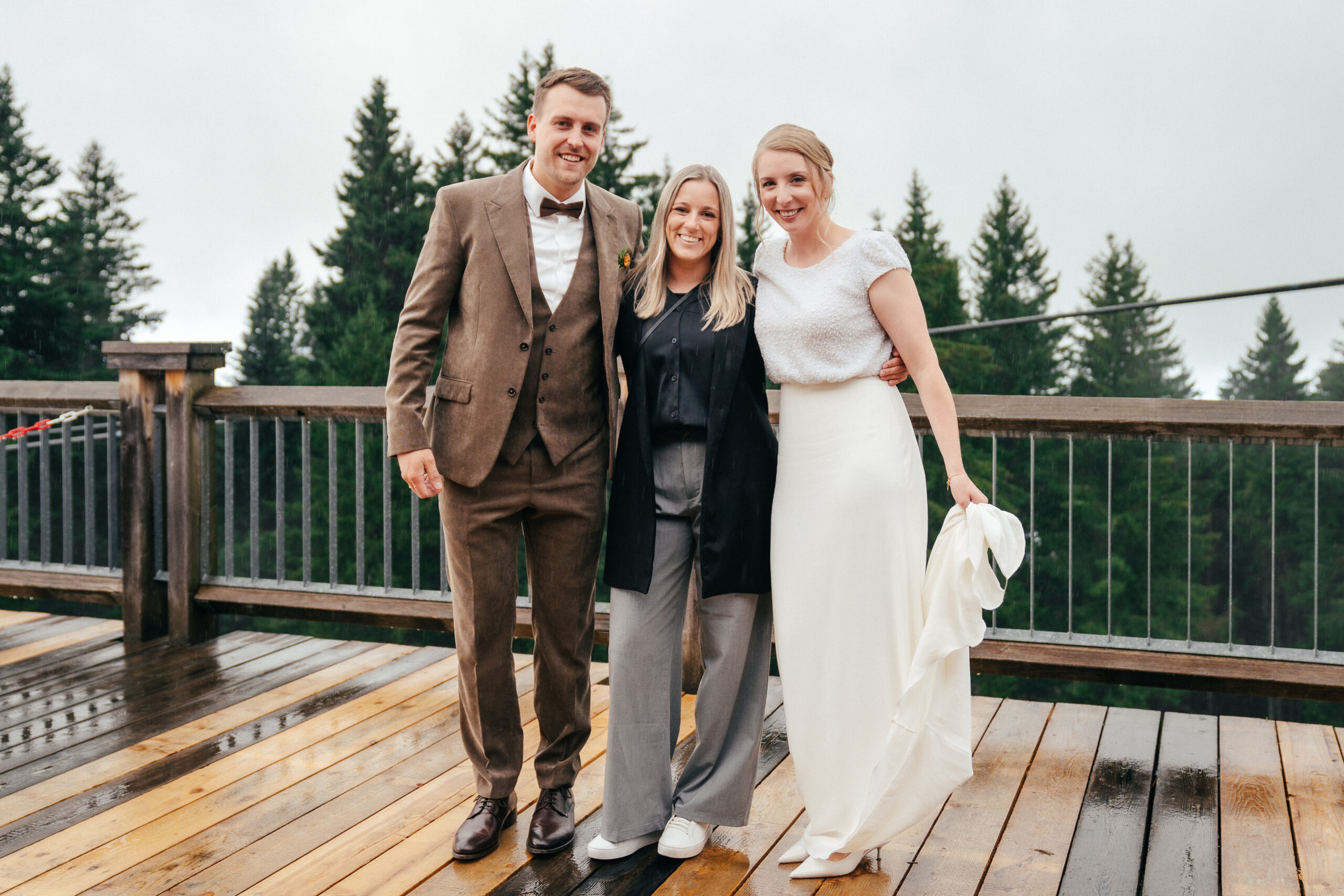 Drei Personen, darunter Braut und Bräutigam, lächeln und posieren zusammen auf einer nassen Holzterrasse mit Bäumen im Hintergrund und fangen den Charme eines Hochzeitstages ein, der von der natürlichen Schönheit Südtirols inspiriert ist.