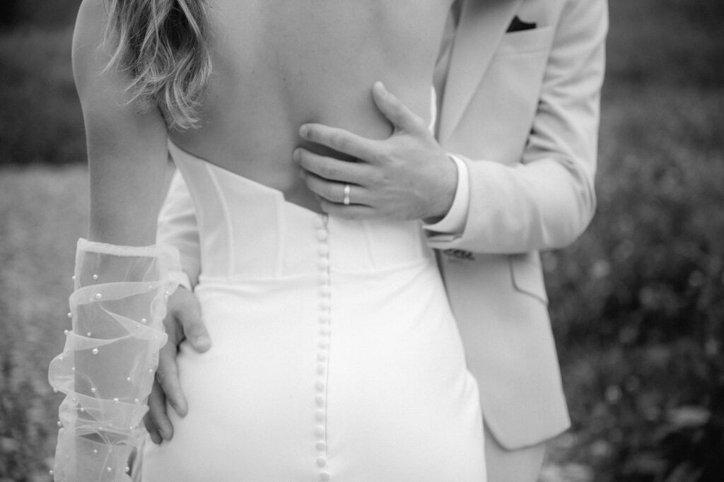 Ein Bräutigam umarmt eine Braut in einem trägerlosen Kleid mit Knöpfen am Rücken; ihr durchsichtiger Handschuh ist während ihres Hochzeitswochenendes in Südtirol zu sehen.
