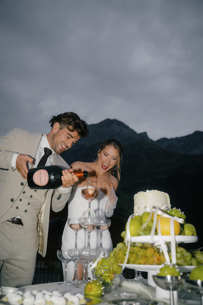 Ein Mann schenkt Champagner in Gläser ein, während eine Frau bei einer Feier im Freien in der Bodenseeregion lacht, während sich im Hintergrund majestätische Berge erheben.