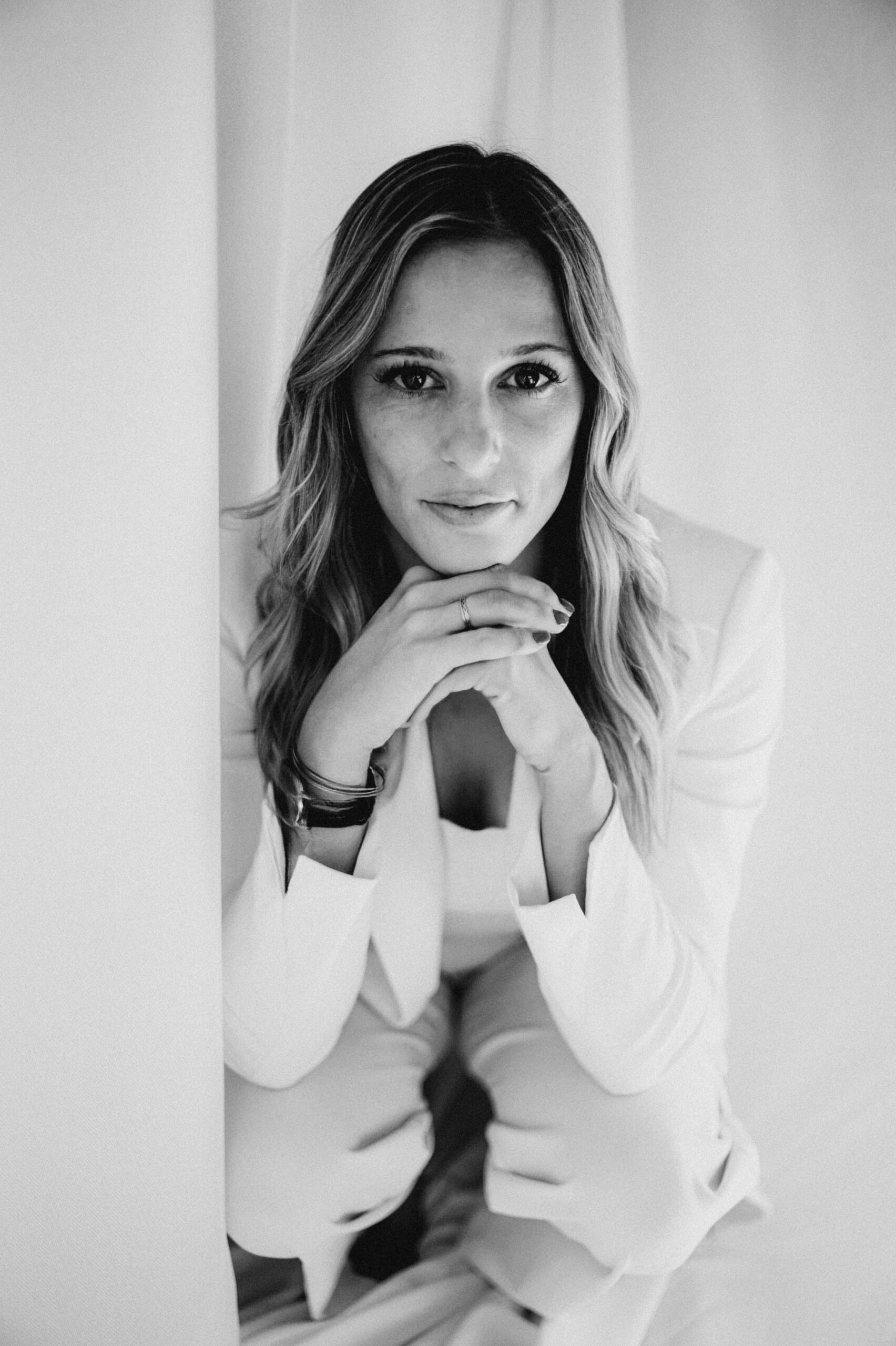 Schwarzweiß-Porträt einer Frau im hellen Anzug, die mit den Händen unter dem Kinn sitzend in die Kamera schaut - aufgenommen während eines Hochzeitswochenendes in der malerischen Bodensee-Region.