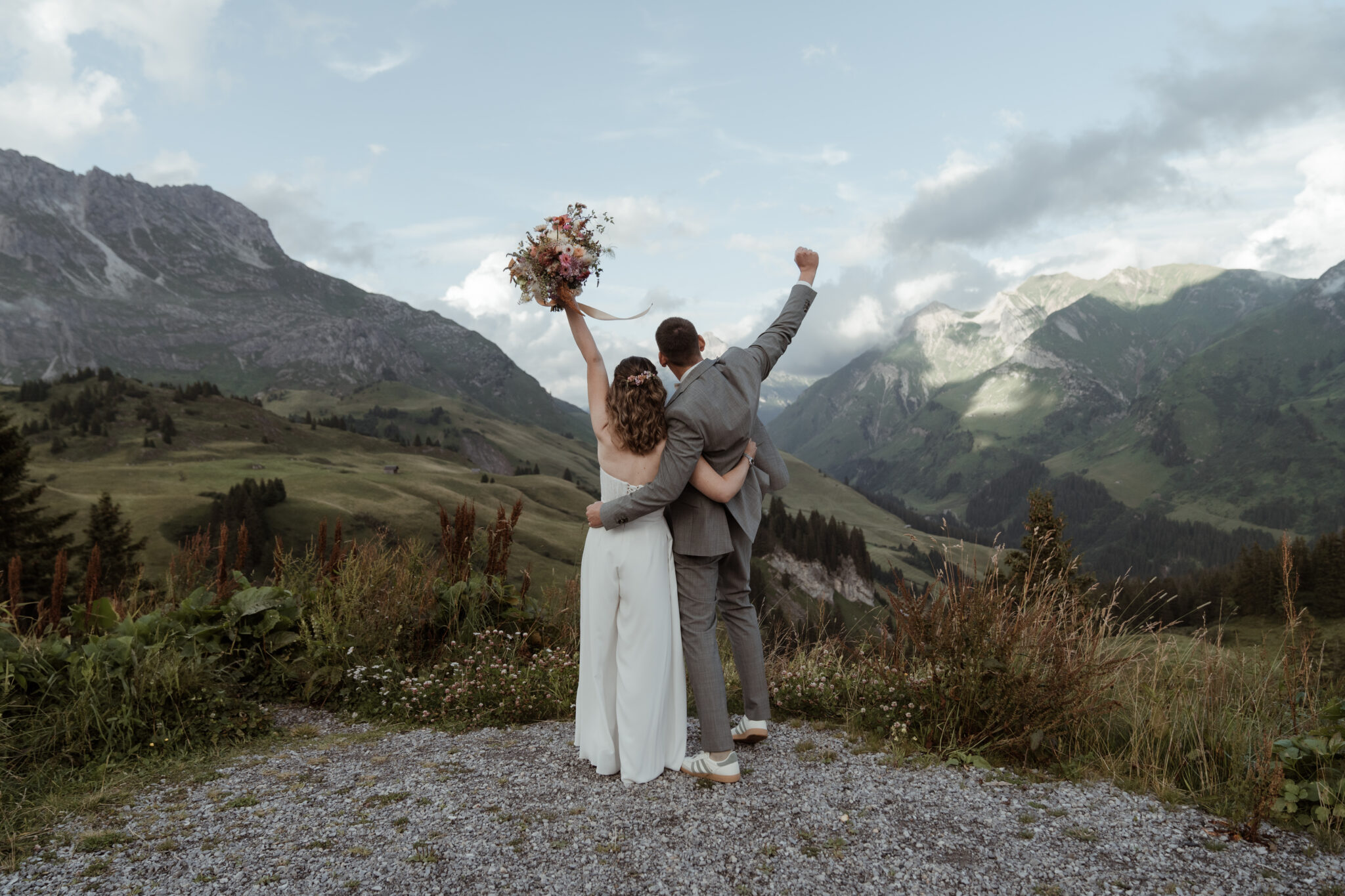 Braut und Bräutigam stehen Arm in Arm und feiern mit erhobenen Armen vor der atemberaubenden Südtiroler Bergkulisse während ihres Hochzeitswochenendes.