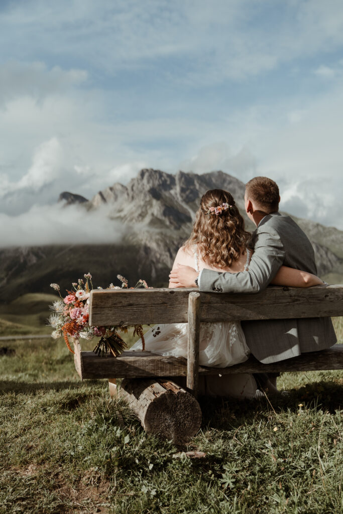 A couple sits on a bench, hugging each other, with a view of the mountains in the heart of South Tyrol and a bouquet of flowers next to them.