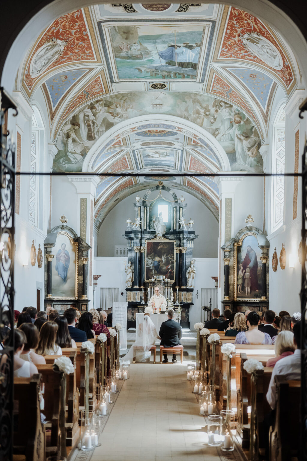 Braut und Bräutigam sitzen während einer Hochzeitszeremonie in einer geschmückten Kirche in Südtirol vor dem Altar, während die Gäste zusehen.