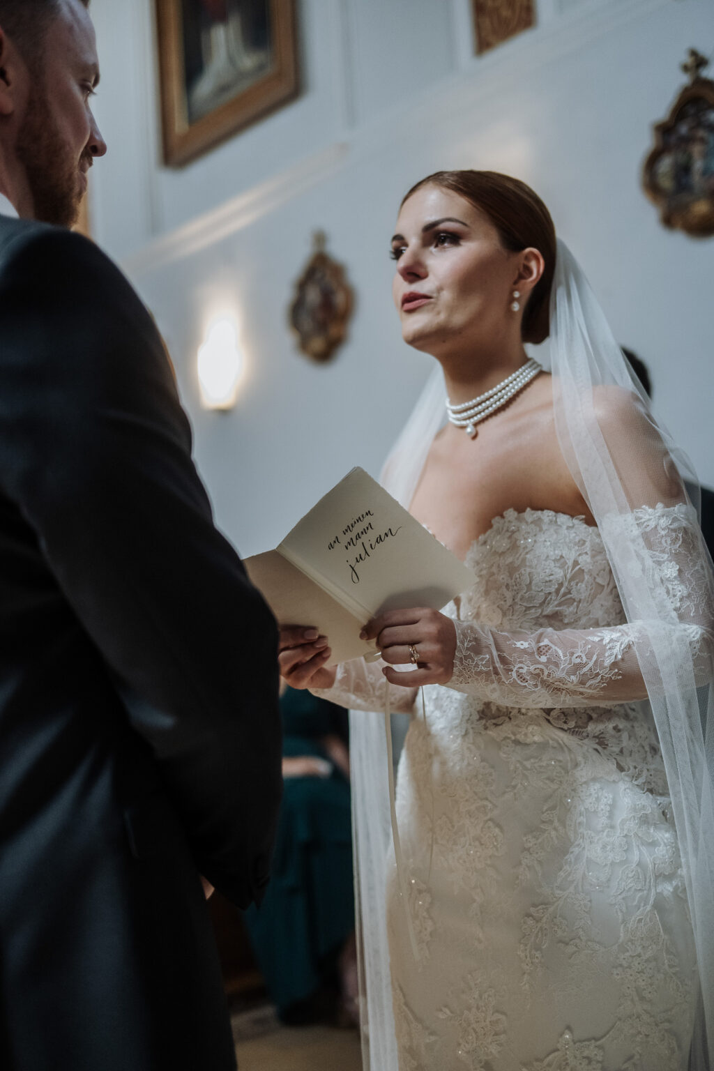 Die Braut im Spitzenkleid liest ihrem Bräutigam das Eheversprechen während einer romantischen Hochzeitszeremonie in einer wunderschön dekorierten Halle in der Bodensee-Region.