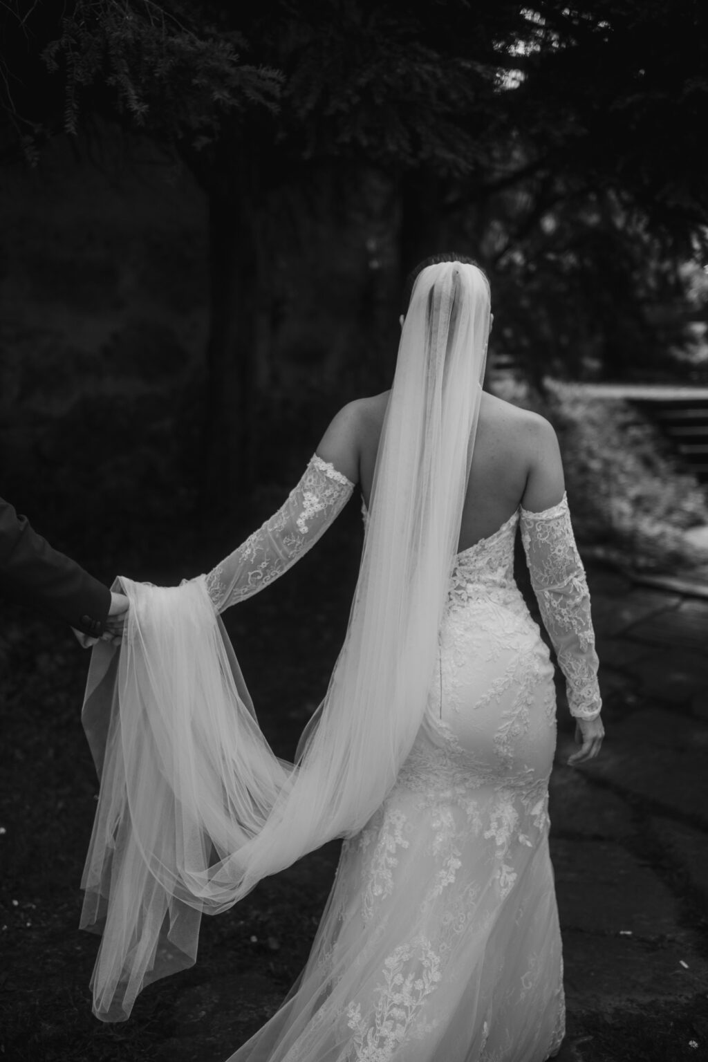 Eine Braut in einem Spitzenkleid und mit langem Schleier geht in der Schweiz im Freien spazieren und hält mit jemandem, der nicht im Bild ist, Händchen - ein schöner Moment für ein Hochzeitswochenende.