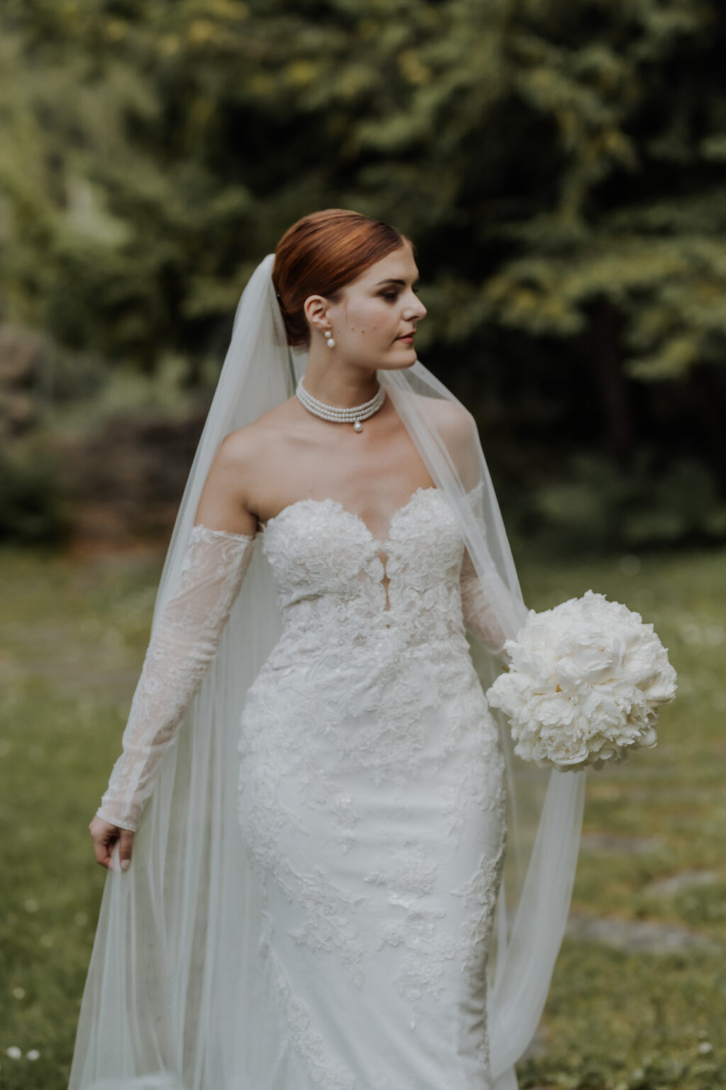 Braut im Spitzenhochzeitskleid mit Schleier und weißem Blumenstrauß im Freien mit üppigem Grün in der schönen Bodenseeregion in Österreich während ihres Hochzeitswochenendes.