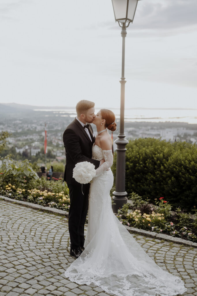 Braut und Bräutigam umarmen und küssen sich im Freien vor einer malerischen Kulisse und einer Straßenlaterne im Hintergrund, um den Zauber ihres Hochzeitswochenendes in der malerischen Bodenseeregion festzuhalten.