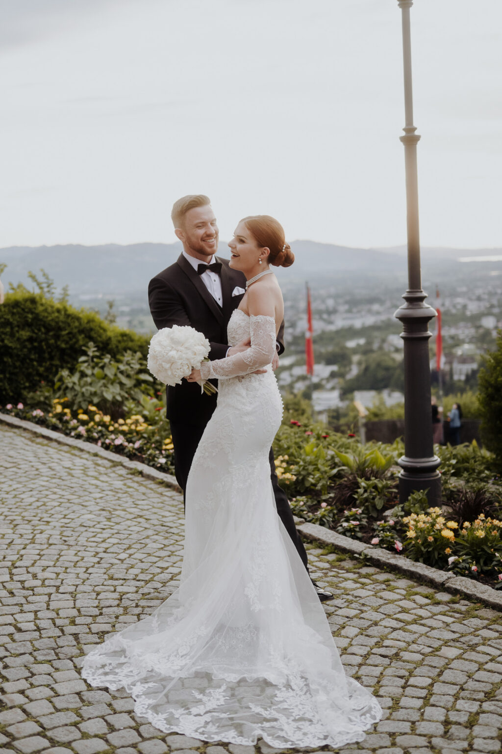 Braut und Bräutigam lächelnd und umarmend im Freien auf einem Kopfsteinpflasterweg, mit einem malerischen Blick auf die Stadt im Hintergrund, um einen romantischen Moment in der charmanten Bodensee-Region einzufangen.