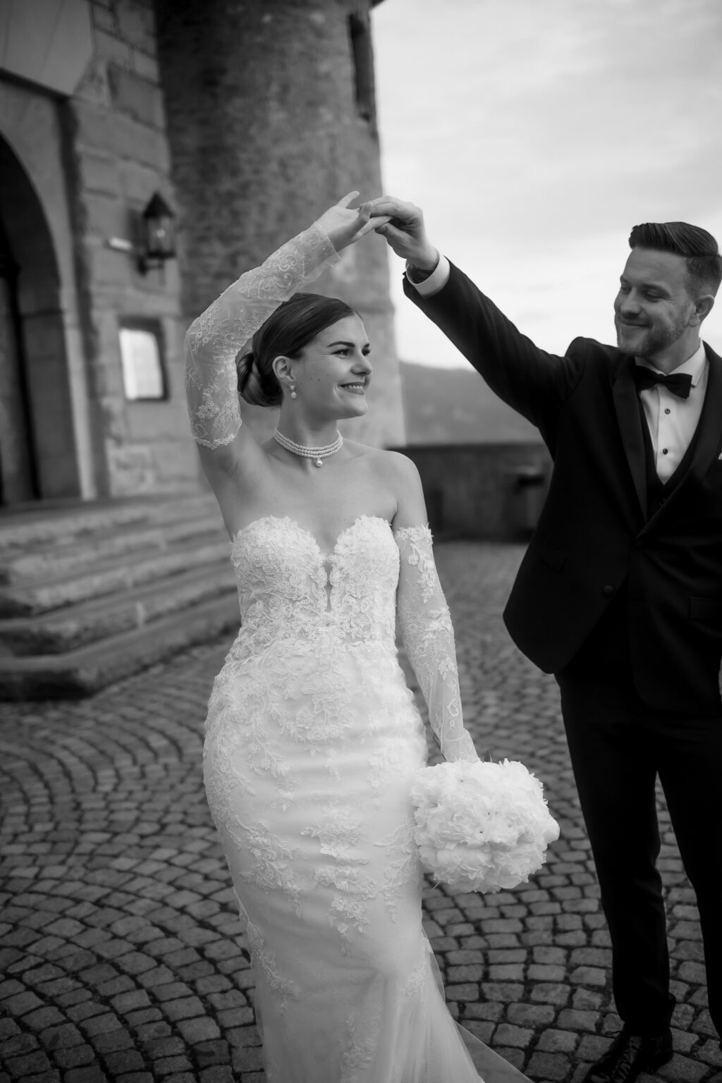 Ein Brautpaar tanzt im Freien auf einem Kopfsteinpflasterweg in Südtirol, lächelnd und händchenhaltend während ihres romantischen Hochzeitswochenendes.
