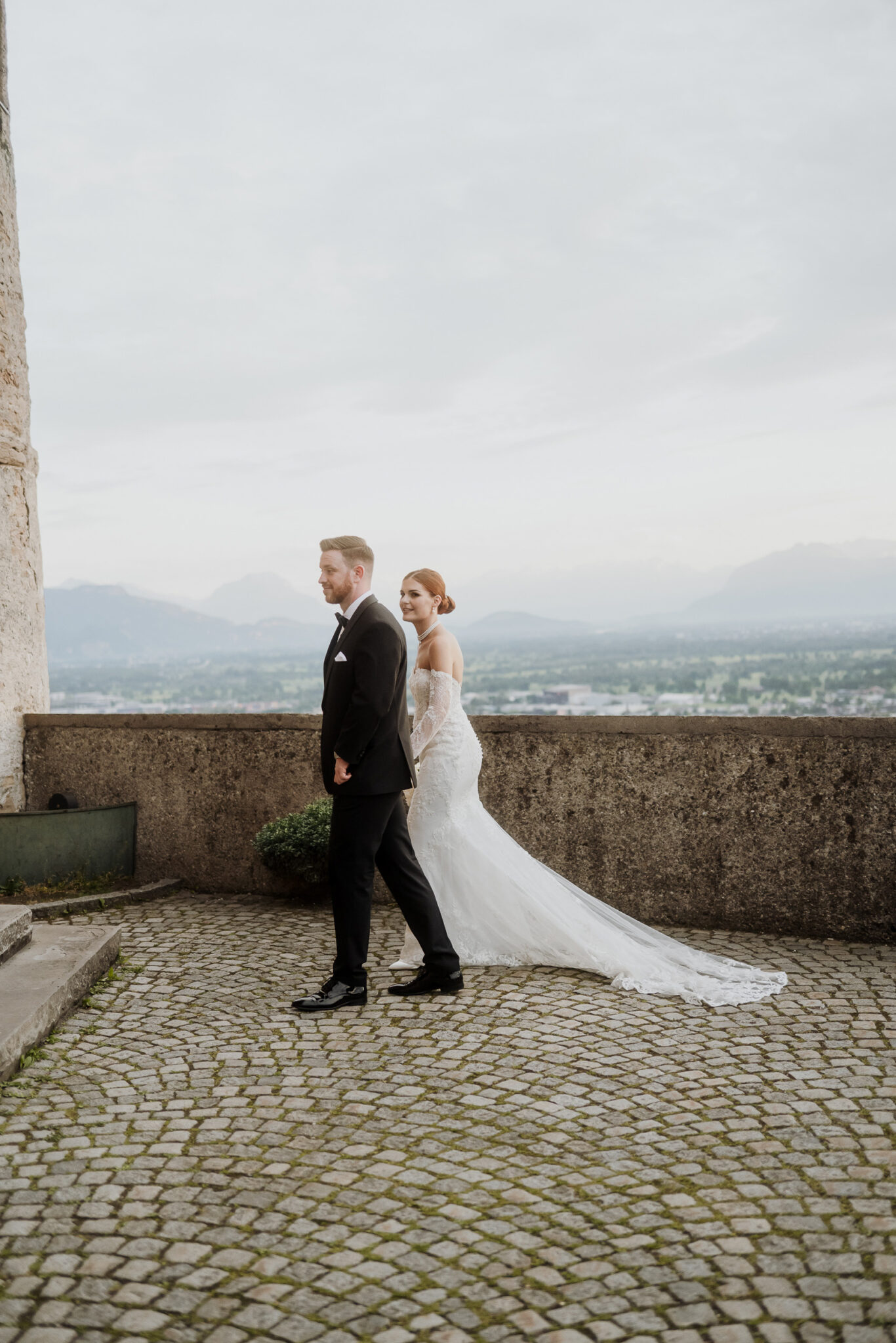 A bride in a white dress walks behind a groom in a suit on a stone terrace, while the majestic mountains of the Lake Constance region rise up in the background.