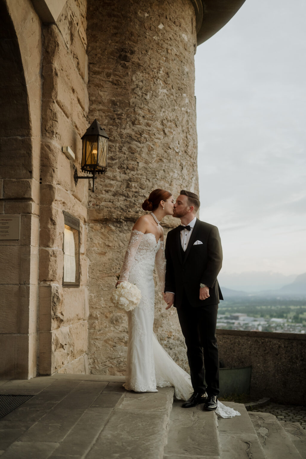 Braut und Bräutigam küssen sich vor einem Steingebäude in Südtirol, halten sich an den Händen und halten einen Blumenstrauß, vor einem malerischen Hintergrund, der die Romantik ihres Hochzeitswochenendes einfängt.