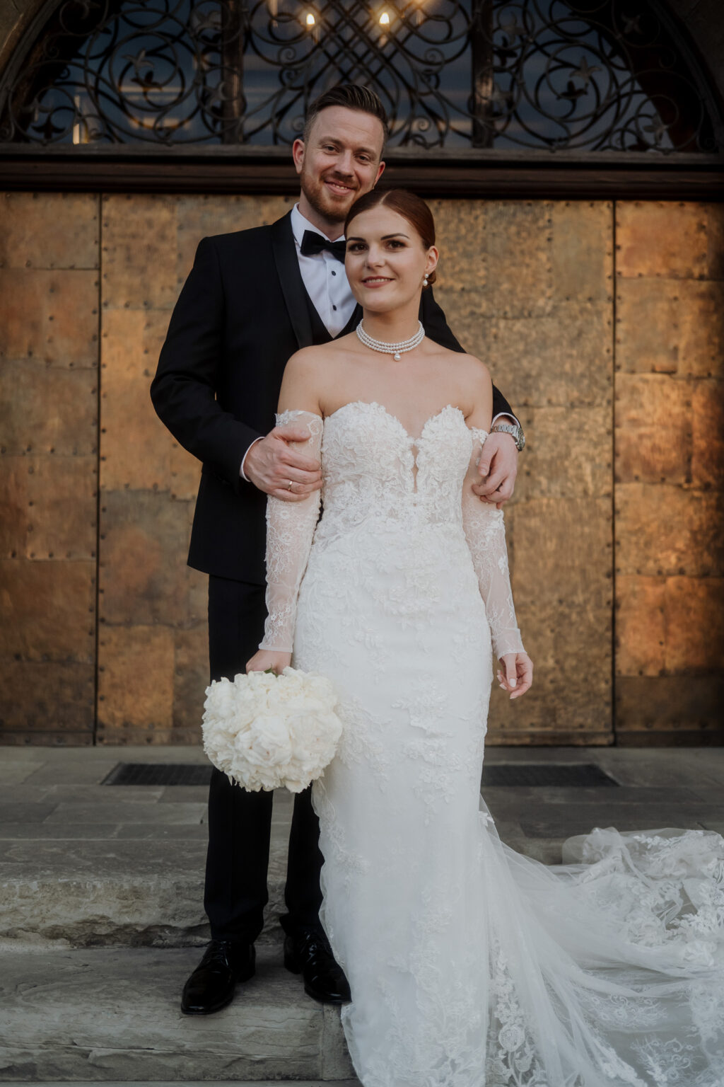 Eine Braut und ein Bräutigam in förmlicher Hochzeitskleidung posieren gemeinsam im Freien vor verschnörkelten Bronzetüren und fangen damit die zeitlose Eleganz von Feierlichkeiten in Österreich und Südtirol ein.