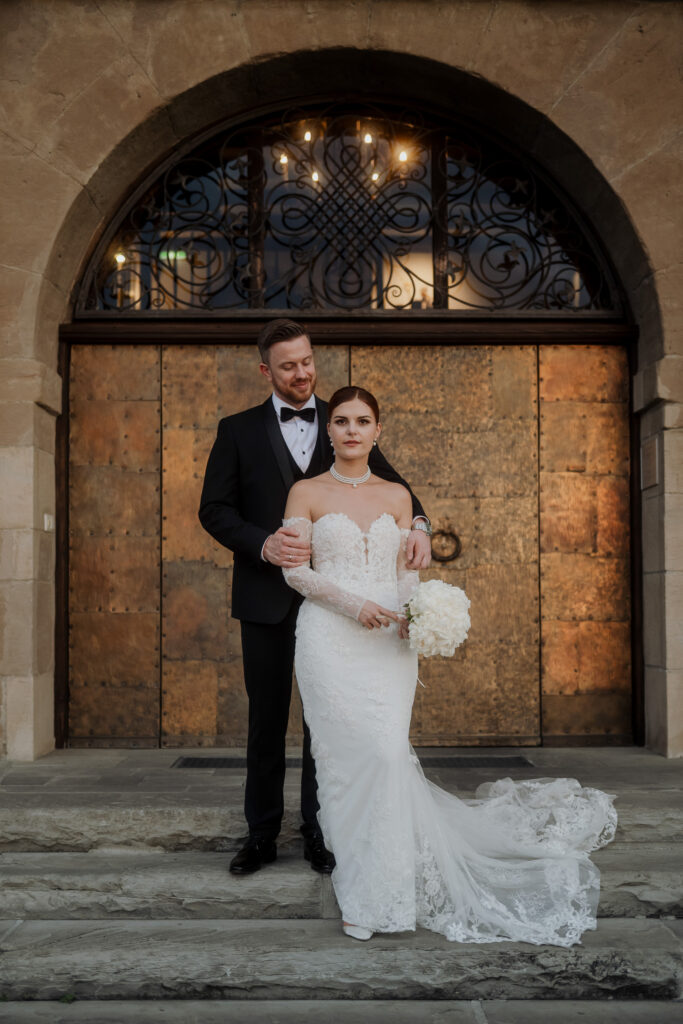 Die Braut in einem weißen Kleid hält Blumen, während der Bräutigam im Smoking hinter ihr auf den Steinstufen vor den verzierten Türen steht. Ein eleganter Moment, der von Hochzeiten in Österreich und Südtirol inspiriert ist.