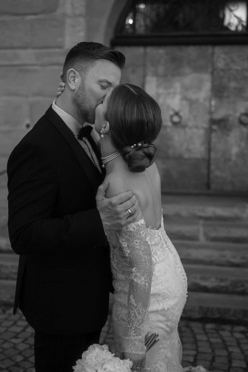 Eine Braut und ein Bräutigam küssen sich im Freien in der malerischen Bodensee-Region, die Braut hält einen Blumenstrauß, beide sind in formelle Hochzeitskleidung gekleidet.