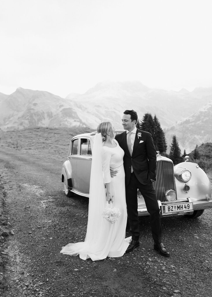 Bride and groom stand next to a vintage car on a mountain road in the Lake Constance region and look at each other, black and white photo.