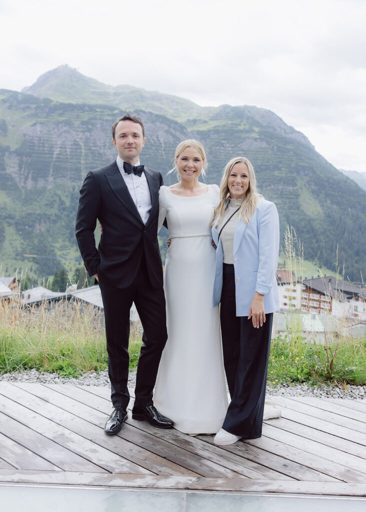 Three people pose outdoors in the Lake Constance region; one wears a tuxedo, one a white dress and one a light-colored blazer, with the mountains in the background - capturing the joy of a wedding weekend in Austria.