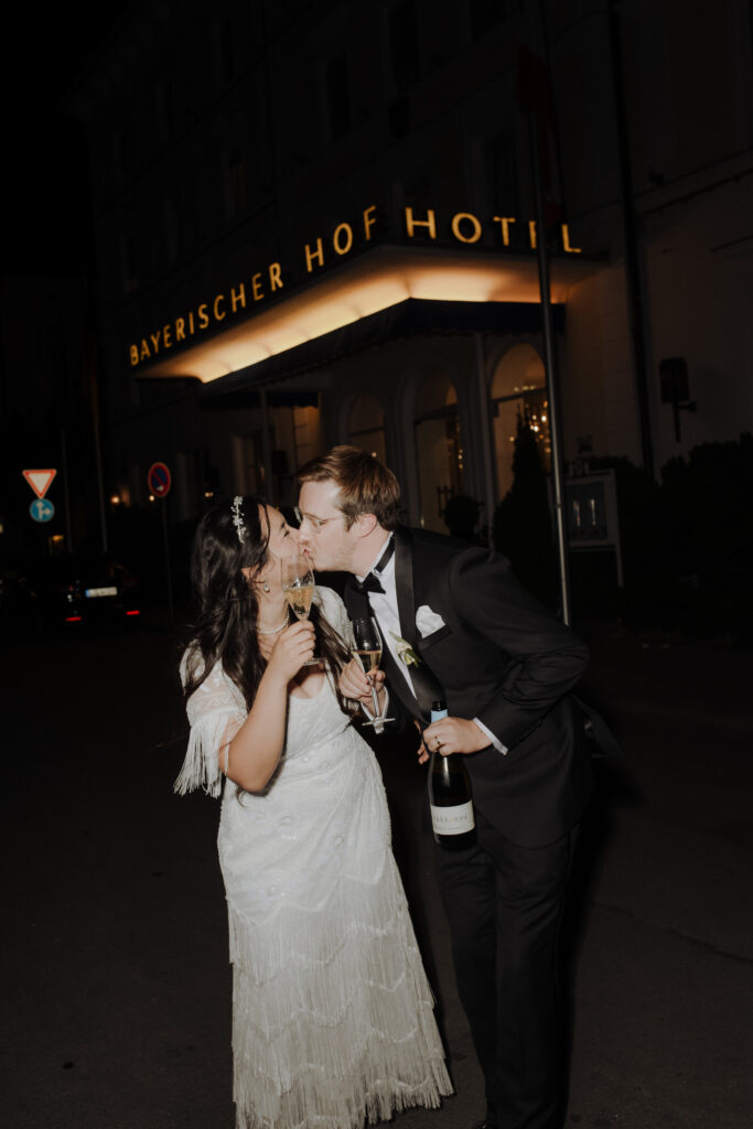 A newly married couple kiss with a glass of champagne in their hands in front of the Hotel Bayerischer Hof and celebrate their love in the heart of the Lake Constance region.
