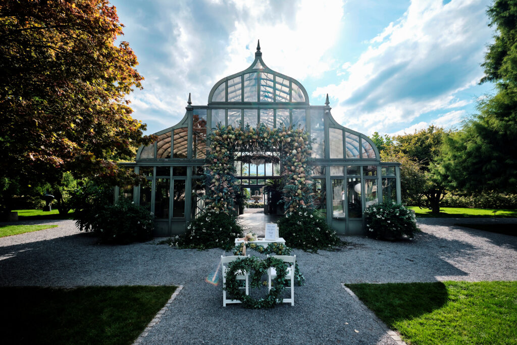 A glass greenhouse decorated with flowers, a white table and chairs set outside on a sunny day evoke the charming atmosphere of the Lake Constance region.
