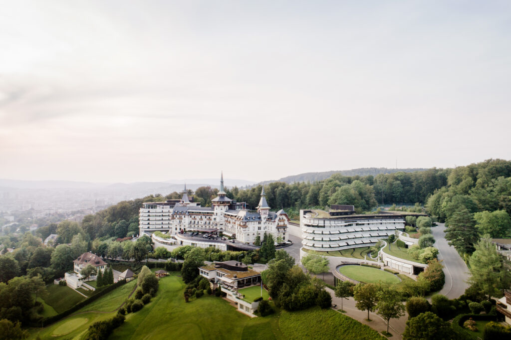 A large, luxurious hotel complex surrounded by greenery and hills, with a mix of modern and historic buildings - an ideal setting for an unforgettable wedding weekend inspired by the charm of Switzerland and Austria.