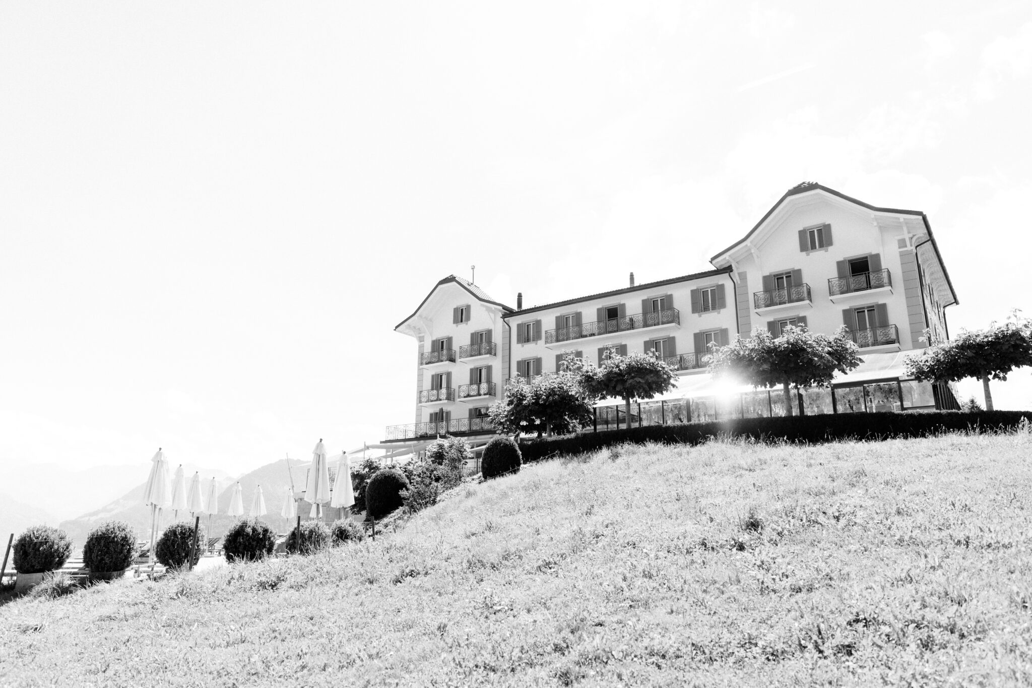 Black and white photo of a large hotel on a grassy hill in South Tyrol, with trees and parasols on the terrace in front.