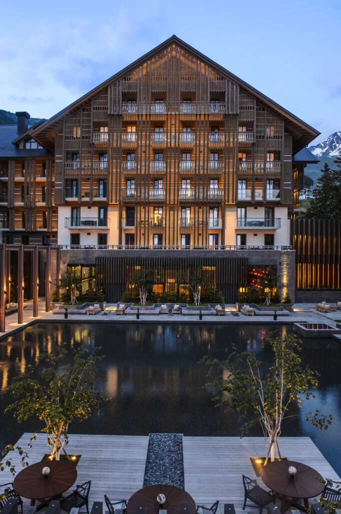 Modernes Holzhotel mit Balkonen, die in der Abenddämmerung beleuchtet werden, mit Blick auf einen ruhigen Pool und einen Sitzbereich im Freien.