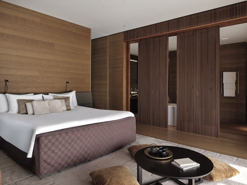 Modernes Hotelzimmer mit Holzvertäfelung, einem ordentlich gemachten Bett, Schiebetüren und einem niedrigen Tisch mit Kissen.