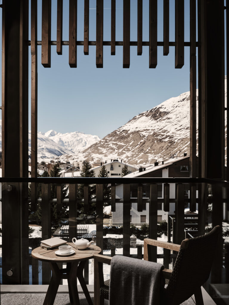 Ein Balkon mit einem gedeckten Kaffeetisch bietet einen Blick auf die schneebedeckten Berge und die Dächer des Dorfes unter blauem Himmel.
