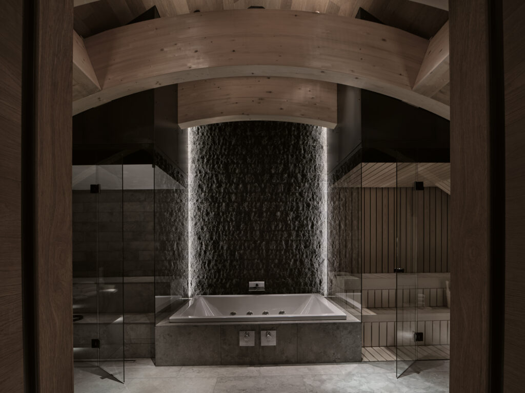 Luxuriöses, modernes Badezimmer mit eingelassener Badewanne, Steinwand, Holzbalken und glasüberdachter Sauna.